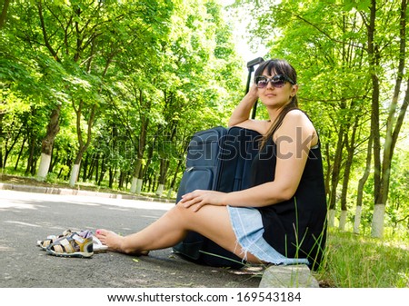 Attractive barefoot woman going on summer vacation sitting on the curb at the side of a rural tree-lined road waiting with her suitcase for a lift