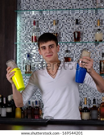 Young Caucasian barman smiling while shaking two cocktail shakers behind the bar with shelves with bottles of alcoholic beverages in the background