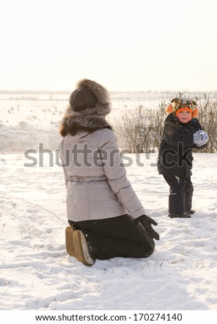 Small boy taking aim and throwing a snowball at his mother as she kneels down in the winter snow as they play together enjoying the winter weather