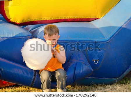 Cute small boy enjoying a stick of candy floss eating it while sitting on a plastic jumping castle at a funfair