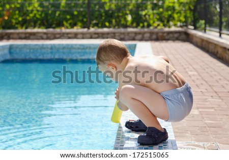 Young boy playing at the edge of a swimming pool in his costume and shoes as he prepares to put something in the cool blue water on a hot sunny day