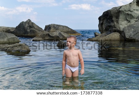 Small boy paddling in his swimsuit in a rocky tidal pool at the seaside standing looking at the camera with his hands on his hips