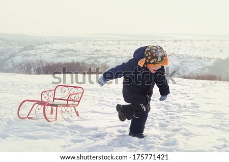 Little boy running through snow dragging a colourful orange toboggan behind him as he enjoys his freedom in the winter countryside