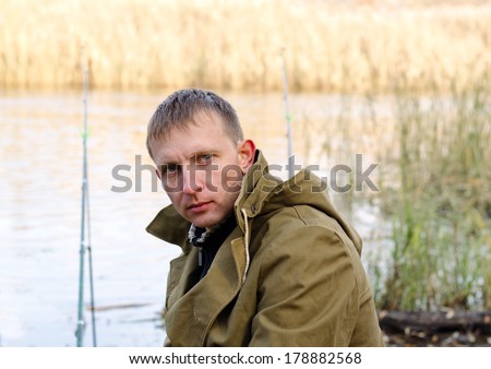 Middle-aged fisherman fishing at a lake dressed in a rugged sports jacket turning to look at the camera with a serious expression with his rods in the background