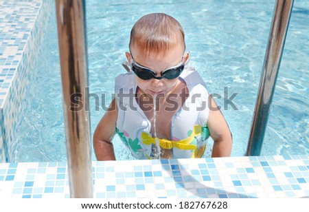 Little boy spitting out a mouthful of water as he hangs onto the steps at the side of a swimming pool in his goggles and flotation jacket