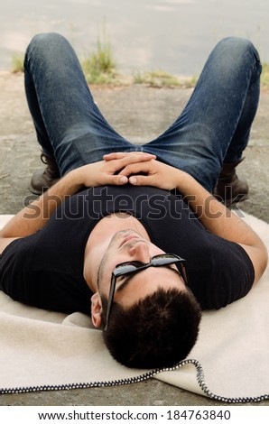 Handsome young man wearing sunglasses lying on his back on a rug relaxing in the sun with his hands folded comfortably on his chest as he unwinds