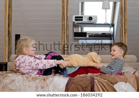 Brother and sister having a tug of war as they lie together on a bed pulling in opposite directions on a large soft stuffed toy dog