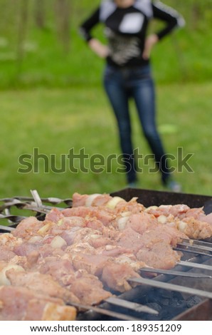 Meat kebabs cooking over a summer BBQ at a rural campsite with a woman standing in the background with focus to the meat