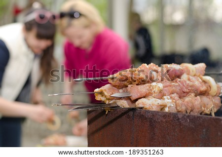 Kebabs ready for cooking standing piled on the grill on an outdoor BBQ as two women continue to prepare more food in the background, focus to the kebabs