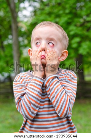 Little boy pulling a scary expression pulling down on his cheeks with his hands to distort his eyes as he plays outdoors in a garden