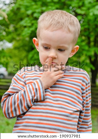 Candid image of a thoughtful little boy standing picking his nose outdoors in a garden or park