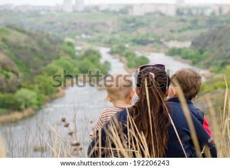 Mother with two small boys enjoying a day in nature sitting in the tall grass on the summit of a hill overlooking a picturesque valley with a wide river with islands, view from behind