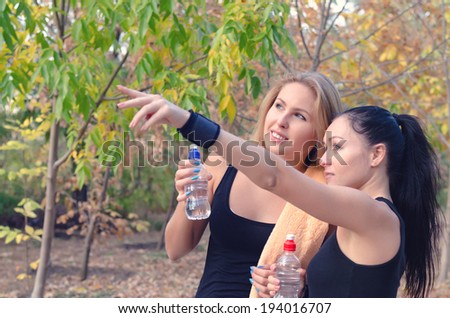 Two fit young women athletes drinking bottled water as they take a break from their training workout as one points to something in the distance