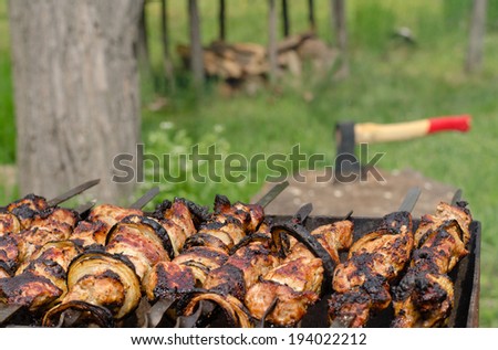Kebabs sizzling on a grill over a hot BBQ fire with a chopper on a chopping block for cutting the wood and fuel for the fire is visible in the background