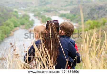 Mother with two small boys enjoying a day in nature sitting in the tall grass on the summit of a hill overlooking a picturesque valley with a wide river with islands, view from behind