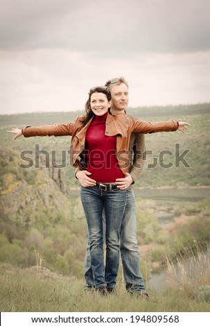 Attractive happy couple posing in the wilderness standing on a grassy hill in a close embrace with the attractive young woman smiling happily as she playfully holds her arms extended sideways