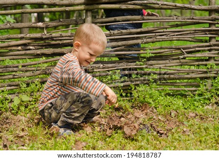 Little boy starting a fire outdoors kneeling down on the ground with his box of matches setting fir to a pile of leaves and twigs watched from behind a rustic wooden fence by a friend