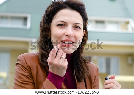 Beautiful young woman applying lipstick to her lips outside a house as she refreshes her makeup during the day, close up of her face