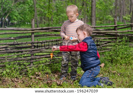 Two small boys lighting a fire in woodland setting fire to a pile of leaves and twigs in the grass as they enjoy a day camping in nature