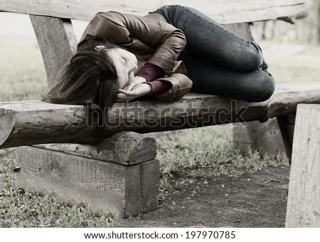 Monochrome image of a woman lying curled up sleeping on a rustic wooden park bench, conceptual of homelessness, exhaustion and loneliness