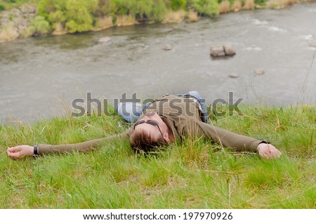 Young man relaxing enjoying nature lying on his back on a grassy hill overlooking a river valley with his arms outspread in the sunshine