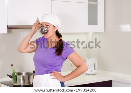 Attractive female chef in a white toque and apron sampling the recipe in the kitchen sipping from a ladle with a thoughtful expression