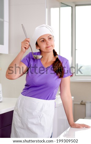 Attractive female chef in a white toque and apron sampling the recipe in the kitchen sipping from a ladle with a thoughtful expression