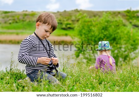 Young boy playing with a pair of binoculars as he sits at the edge of a rural lake with a fair haired little girl visible behind him in the long grass