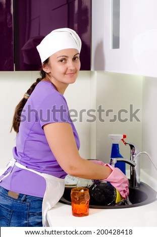 Smiling woman cook in a white apron and hat cleaning dishes at the kitchen sink after cooking a meal