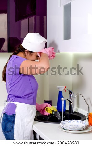 Tired or flustered female cook in a white apron and cap wiping her brow with her gloved hand as she does washing up at the sink