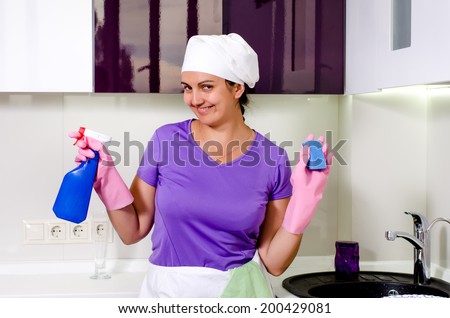 Cute happy playful housewife wearing a white cap over her long hair holding up a spray bottle and sponge as she grins at the camera