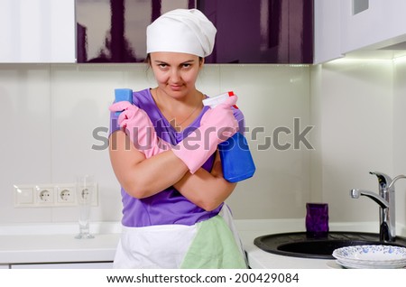 Cute happy playful housewife wearing a white cap over her long hair holding up a spray bottle and sponge as she grins at the camera