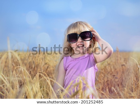 Adorable little blond girl with her hair tied up in pigtails posing in huge fashionable sunglasses in a golden wheat field laughing and waving