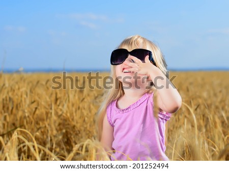 Cute little blond girl playing in a wheat field wearing a pair of large fashionable sunglasses looking up into the sun and laughing