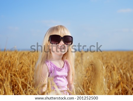 Cute little blond girl playing in a wheat field wearing a pair of large fashionable sunglasses looking up into the sun and laughing
