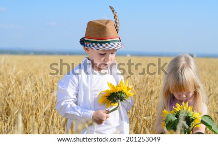 Little girl and boy in a trendy fashionable hat with a feather standing alongside a field of wheat examining fresh bright yellow sunflowers as they learn about nature