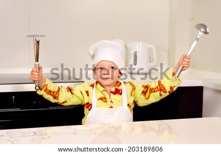 Cute boy chef in a white toque and apron displaying his utensils brandishing a ladle and potato masher in the air before starting to prepare dinner