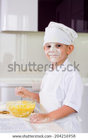 Cute little boy in a chefs uniform baking a cake in the kitchen adding ingredients from the recipe into his mixing bowl
