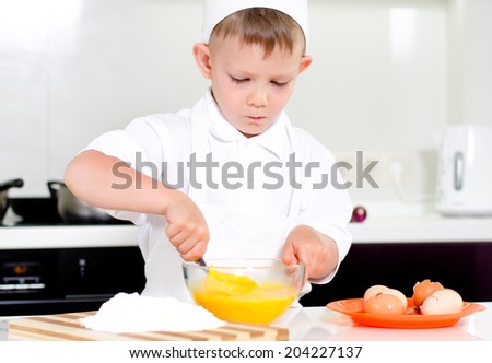 Young boy in a chefs uniform and toque baking whipping eggs in a mixing bowl as he bakes a cake in the kitchen