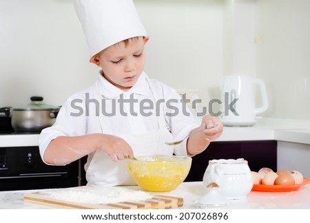 Little boy in a chefs uniform baking in the kitchen spreading flour on his wooden chopping board before rolling out his dough from the mixing bowl