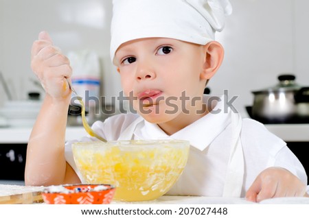 Cute little chef in a white toque and apron tasting his cooking as he mixes ingredients in a bowl looking thoughtfully into the air