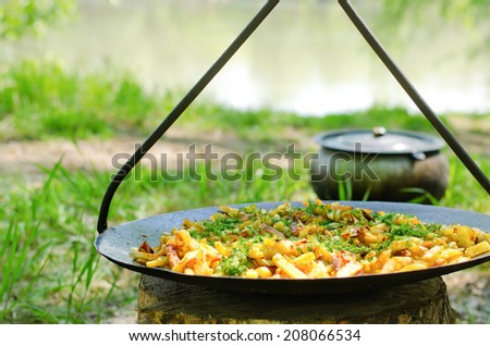 Savory potato wedges or chips and herbs cooking in oil on a barbecue at a campsite alongside a lake with an iron cooking pot visible beyond