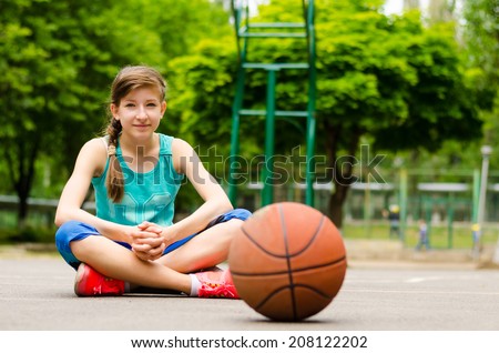 Young female basketball player sitting cross-legged on the outdoor court surrounded by leafy green trees with the ball waiting for play
