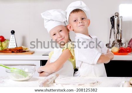 Two cute proud young chefs with a little boy and girl standing back to back in the kitchen in their white uniforms and toques with folded arms smiling at the camera