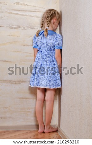 Little girl with her blond hair in braids standing in the corner facing the wall sulking or in punishment for a wrongdoing