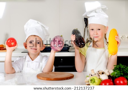 Two smiling young children, a boy and girl wearing white chefs toques and aprons, standing in the kitchen holding up fresh vegetables to be used as ingredients in the meal they are about to cook