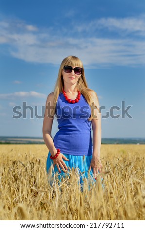 Attractive blond woman wearing sunglasses and a summer top standing enjoying the sun in a wheat field with her arms outstretched and head tilted to the sun with a smile