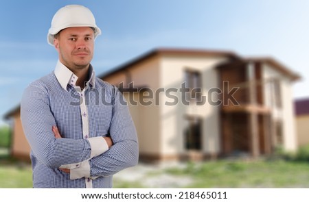 Architect, builder or structural engineer wearing a hardhat standing with folded arms in front of a newly constructed house
