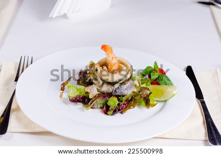 Delicious Main Dish of Fish and Shrimp Meat and Veggies Combination in Stylish Plating on White Round Plate with Fork and Knife on Sides.