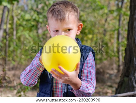 Small boy blowing up a colorful yellow party balloon with a look of concentration as he stands outdoors in the garden